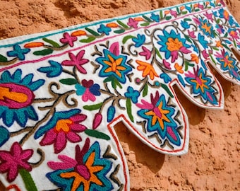 May include: A white fabric valance featuring vibrant, multi-colored floral embroidery in shades of bright pink, turquoise, orange, and green. The bottom edge is cut into a series of pointed, scalloped shapes, each outlined with a thin red thread.