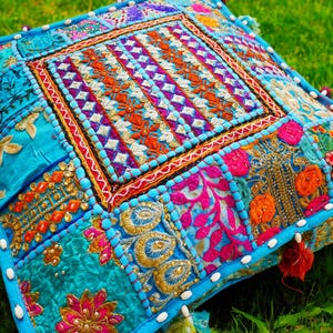 Può includere: Cuscino quadrato decorativo con un design patchwork. Il cuscino presenta un quadrato centrale con dettagli ricamati arancioni, viola e bianchi. Le toppe circostanti sono turchesi, rosa e oro, con bordi perlati e nappe.