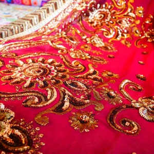 May include: Close-up of a vibrant fuchsia fabric adorned with intricate gold embroidery and sequins. The design features floral and paisley patterns, creating a luxurious and ornate textile. The fabric's texture and embellishments suggest a high-end garment or decorative piece.