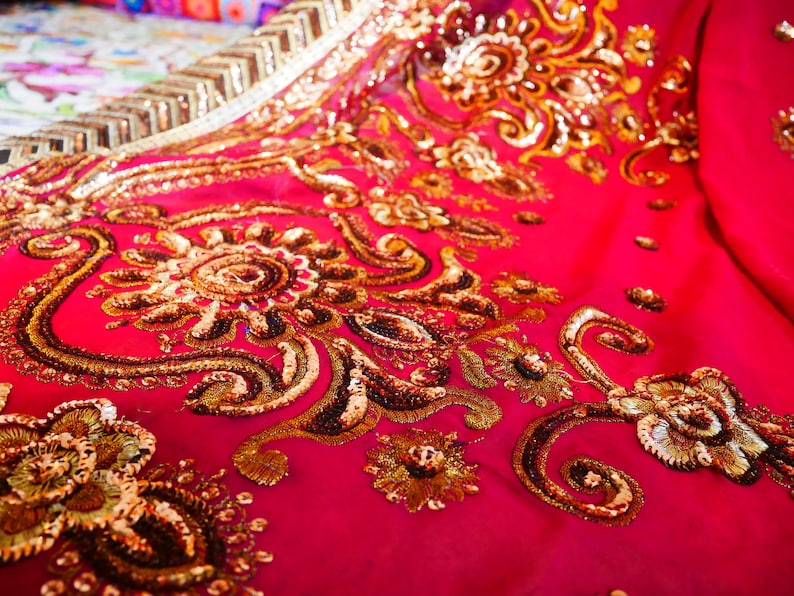 May include: Close-up of a vibrant fuchsia fabric adorned with intricate gold embroidery and sequins. The design features floral and paisley patterns, creating a luxurious and ornate textile. The fabric's texture and embellishments suggest a high-end garment or decorative piece.