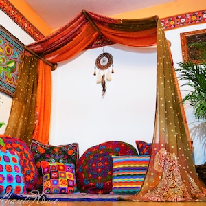 Puede incluir: Una zona de estar de estilo bohemio con almohadas y mantas coloridas. El área está decorada con cortinas transparentes, un atrapasueños y plantas en macetas. El texto "The Shanti Home" es visible en la esquina inferior izquierda de la imagen.