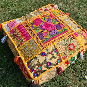 Puede incluir: Un cojín de suelo de patchwork amarillo, rosa y verde con diseños florales bordados. El cojín está hecho de retazos de tela cosidos juntos y tiene un borde con flecos.