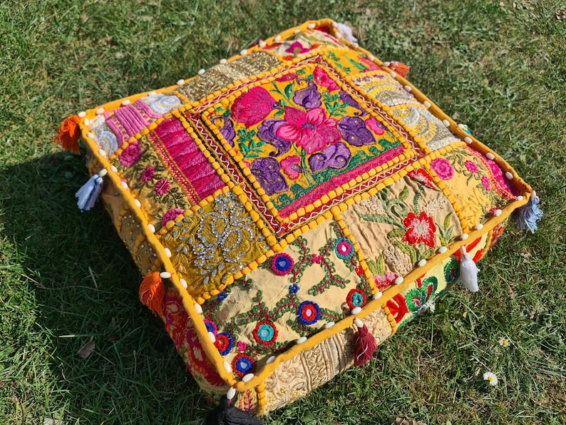 May include: A yellow, pink, and green patchwork floor cushion with embroidered floral designs. The cushion is made of fabric scraps sewn together and has a fringed edge.
