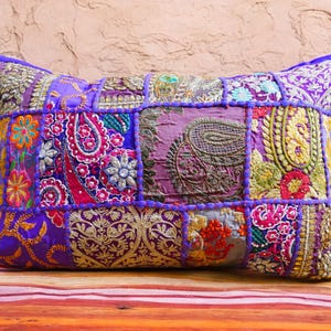 Puede incluir: Cojín rectangular de patchwork con una vibrante mezcla de colores y patrones. El cojín presenta una base morada con cuadrados de tela bordada en tonos rosa, dorado, verde y azul. Los bordes están ribeteados con un borde morado.