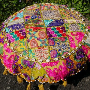 May include: A round, multi-coloured floor cushion with a patchwork design. The cushion is made of fabric with various embroidered patterns and is decorated with white seashells around the edge. The cushion has a fringe trim.
