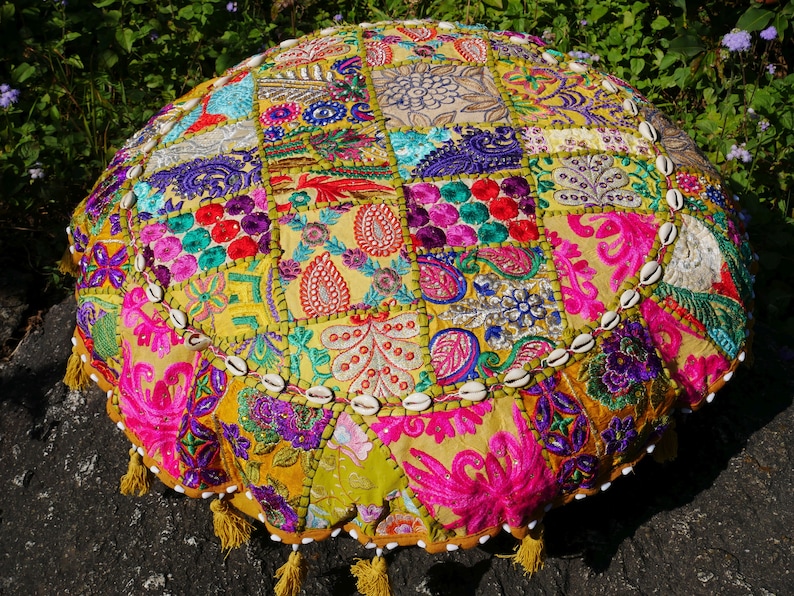 May include: A round, multi-coloured floor cushion with a patchwork design. The cushion is made of fabric with various embroidered patterns and is decorated with white seashells around the edge. The cushion has a fringe trim.