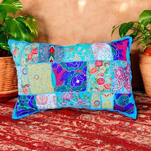 May include: A rectangular patchwork cushion featuring a variety of floral and paisley designs in blue, purple, and pink. The cushion has a bright blue border and is placed on a red patterned rug.