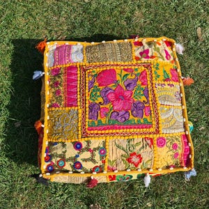 May include: A yellow, square floor cushion with a patchwork design. The cushion is made of different fabrics with floral embroidery in shades of pink, purple, and green. The cushion has a fringe border and tassels.