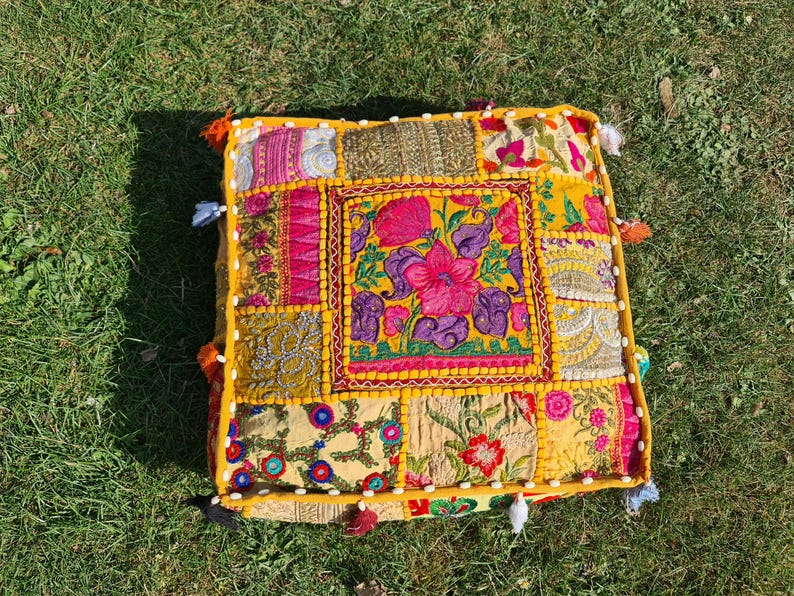 May include: A yellow, square floor cushion with a patchwork design. The cushion is made of different fabrics with floral embroidery in shades of pink, purple, and green. The cushion has a fringe border and tassels.