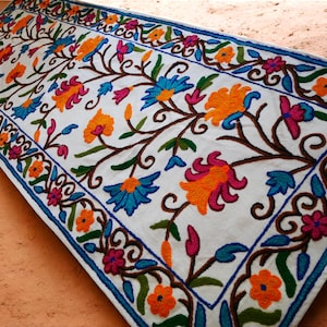 Op de afbeelding: Een wit loperkleed met een geborduurd bloemenontwerp. Het kleed heeft een herhalend patroon van kleurrijke bloemen en ranken, waaronder oranje, roze, blauwe en groene tinten.