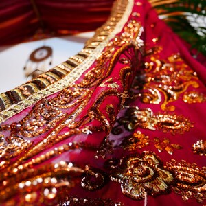 May include: Close-up of a vibrant red fabric with intricate gold embroidery and sequins. The fabric is draped, showcasing detailed patterns and textures. The design includes floral and paisley motifs, creating a luxurious and ornate appearance.