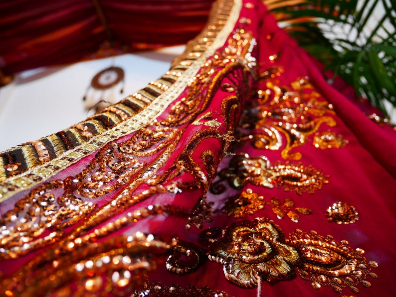 May include: Close-up of a vibrant red fabric with intricate gold embroidery and sequins. The fabric is draped, showcasing detailed patterns and textures. The design includes floral and paisley motifs, creating a luxurious and ornate appearance.