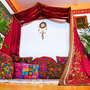 May include: A bohemian-style seating area with a canopy bed draped in deep red fabric. Colorful embroidered pillows in various shapes and sizes are arranged on a rug. A dreamcatcher hangs from the canopy, and a plant adds a touch of greenery.
