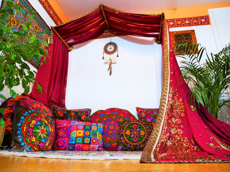 May include: A bohemian-style seating area with a canopy bed draped in deep red fabric. Colorful embroidered pillows in various shapes and sizes are arranged on a rug. A dreamcatcher hangs from the canopy, and a plant adds a touch of greenery.