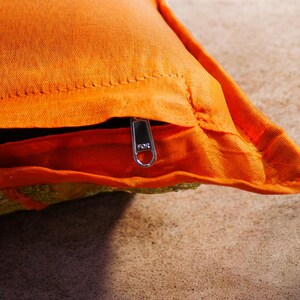 May include: Orange throw pillow with a zipper closure. The pillow has a gold embroidered design on the front.