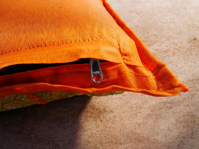 May include: Orange throw pillow with a zipper closure. The pillow has a gold embroidered design on the front.