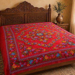 Könnte beinhalten: Eine leuchtend rote Tagesdecke mit einem zentralen Blumenmotiv und einer dekorativen Bordüre. Der Stoff ist mit aufwendigen Stickereien in Blau-, Gelb-, Lila- und Grüntönen verziert, die ein detailliertes Muster auf der gesamten Fläche bilden.