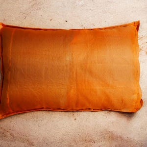 May include: A large, rectangular orange pillow with a smooth, slightly textured surface. The pillow is lying flat on a sandy surface.