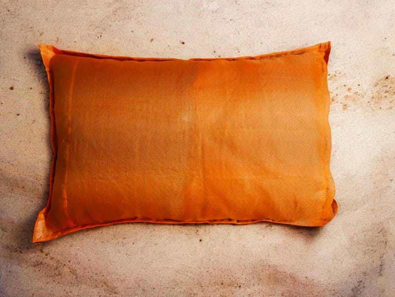 May include: A large, rectangular orange pillow with a smooth, slightly textured surface. The pillow is lying flat on a sandy surface.