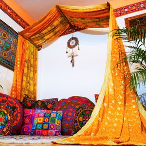 May include: A bohemian-style interior scene featuring a canopy draped with orange and gold fabric. Colorful, embroidered pillows and cushions are arranged on the floor. A dreamcatcher hangs from the canopy, and a tapestry adorns the wall, creating a vibrant and inviting space.