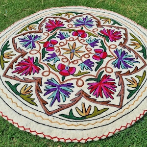 May include: A round, white, embroidered rug with a floral design. The rug features a central motif of a flower with petals in shades of pink, red, and yellow, surrounded by smaller flowers in shades of purple, blue, and green.
