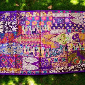 May include: A purple tapestry with a patchwork design featuring various colors and patterns, including gold, pink, and blue. The tapestry is made of fabric and has a fringed edge.