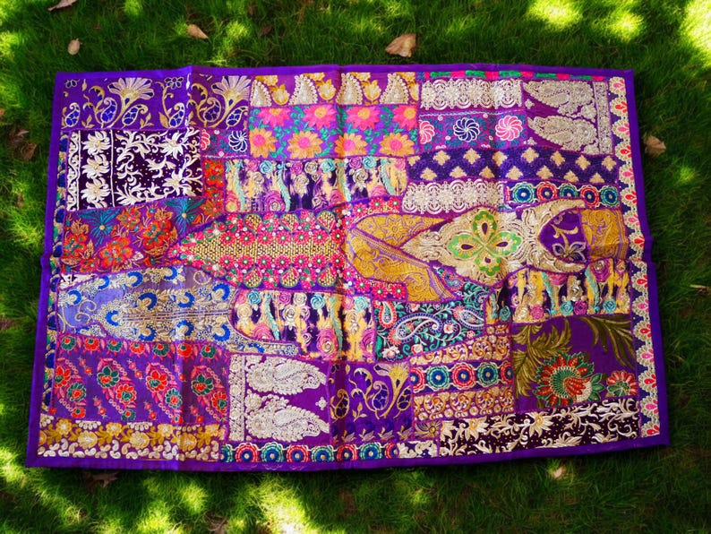 May include: A purple tapestry with a patchwork design featuring various colors and patterns, including gold, pink, and blue. The tapestry is made of fabric and has a fringed edge.