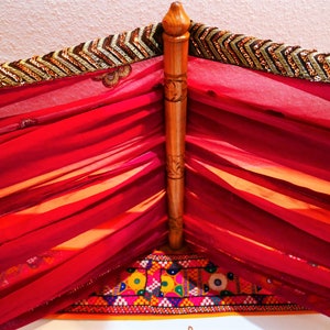 May include: A decorative canopy featuring a wooden frame and draped with sheer, vibrant red fabric. The top edge is adorned with a gold and brown sequined trim. The bottom edge has a colorful embroidered border.