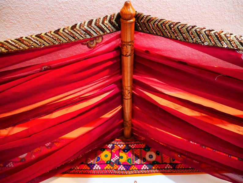 May include: A decorative canopy featuring a wooden frame and draped with sheer, vibrant red fabric. The top edge is adorned with a gold and brown sequined trim. The bottom edge has a colorful embroidered border.