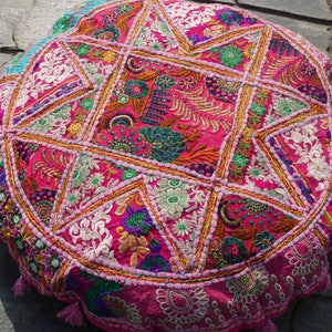 Può includere: Un cuscino da pavimento rotondo rosa e multicolore con ricami e decorazioni intricate. Il cuscino è realizzato in tessuto patchwork e ha una frangia a nappe.