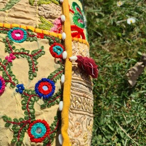 May include: A yellow and beige cushion with colorful embroidery and a red tassel. The cushion is decorated with floral patterns and small mirrors. The cushion is sewn together with white buttons.