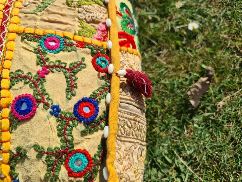 May include: A yellow and beige cushion with colorful embroidery and a red tassel. The cushion is decorated with floral patterns and small mirrors. The cushion is sewn together with white buttons.