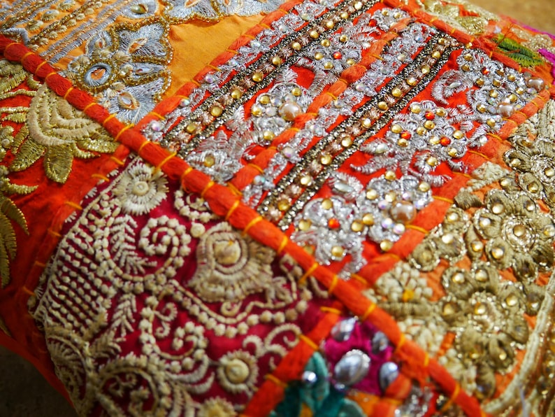 May include: A colorful patchwork cushion cover with intricate embroidery and beaded details. The fabric is a mix of orange, red, and gold, with a variety of patterns and textures.