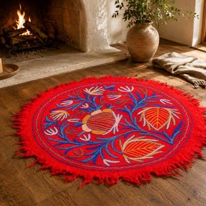 May include: A round, bright red rug featuring a central embroidered floral pattern in shades of blue, orange, and yellow. The rug has a textured, shaggy fringe edge and a circular design that displays stylized botanical motifs across its surface.