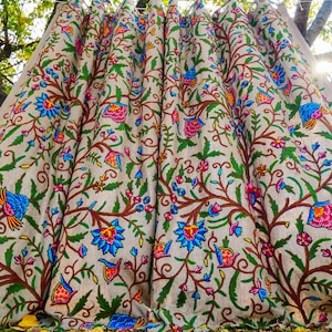 Könnte beinhalten: Ein beiger Stoff mit einem lebendigen Blumenmuster. Das Design zeigt farbenfrohe Blumen, Vögel und Blätter in Blau-, Rosa-, Orange- und Grüntönen, vor dem Hintergrund brauner Äste und Stiele. Der Stoff ist drapiert und zeigt die aufwendige Stickerei.