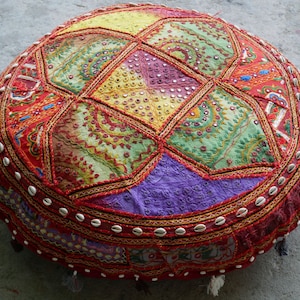May include: Round, patchwork floor cushion in vibrant colors. The cushion features a geometric pattern with red, yellow, green, and purple fabric panels. Embellished with embroidery, small mirrors, and a border of white shells, it adds a bohemian touch.