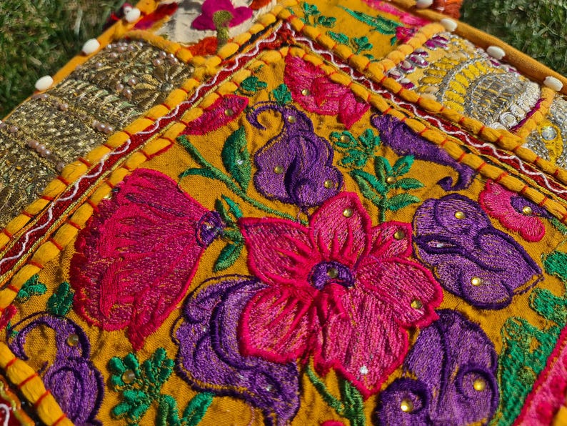 May include: A close-up of a colorful embroidered fabric with a yellow background. The fabric features a floral design with pink, purple, and green accents. The fabric is stitched with a variety of different colored threads.