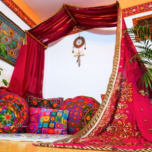 May include: A bohemian-style canopy bed with a burgundy draped fabric and gold trim. The bed is adorned with colorful embroidered pillows and a dreamcatcher. A vibrant tapestry hangs on the wall, complementing the eclectic decor. The scene evokes a cozy, artistic living space.