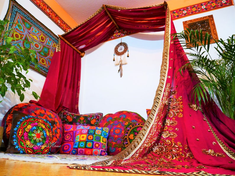 May include: A bohemian-style canopy bed with a burgundy draped fabric and gold trim. The bed is adorned with colorful embroidered pillows and a dreamcatcher. A vibrant tapestry hangs on the wall, complementing the eclectic decor. The scene evokes a cozy, artistic living space.