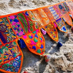 May include: A vibrant, handcrafted textile banner with an orange border. The banner features a patchwork design with embroidered floral and geometric patterns in shades of orange, pink, blue, and green. Decorative tassels hang from the bottom, adding a touch of whimsy.