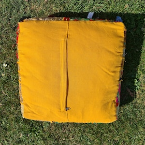 May include: A yellow square cushion with a zipper closure. The cushion is made of a patchwork fabric with colorful floral patterns.