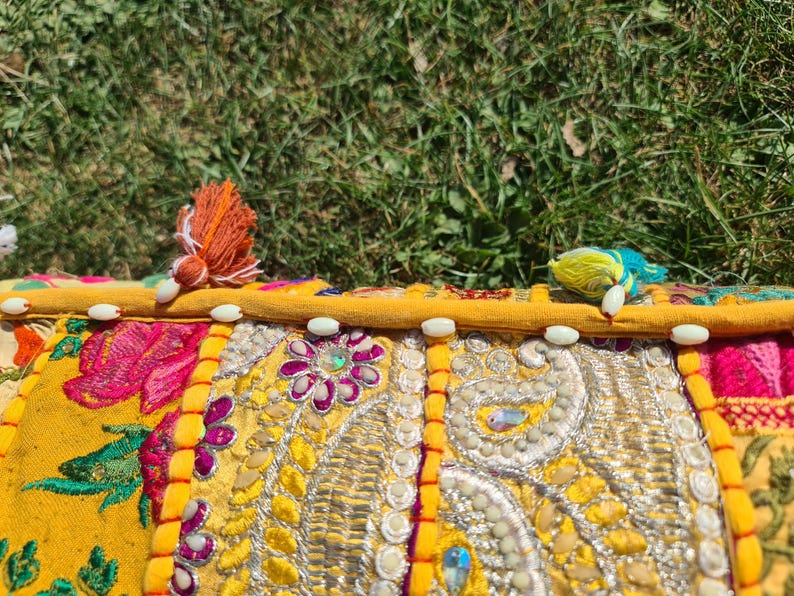 May include: A yellow, pink, and green patchwork cushion with white beads around the edge. The cushion is decorated with embroidered flowers and paisley patterns.
