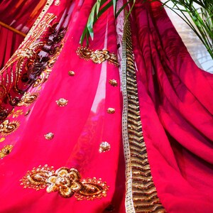 May include: A vibrant fuchsia fabric with intricate gold embroidery and embellishments. The ornate design features floral and geometric patterns. A gold and white trim borders the fabric's edge. The draped material suggests a decorative textile, possibly for a garment or home decor.