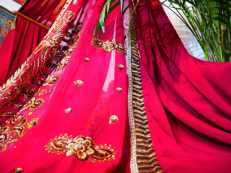 May include: A vibrant fuchsia fabric with intricate gold embroidery and embellishments. The ornate design features floral and geometric patterns. A gold and white trim borders the fabric's edge. The draped material suggests a decorative textile, possibly for a garment or home decor.