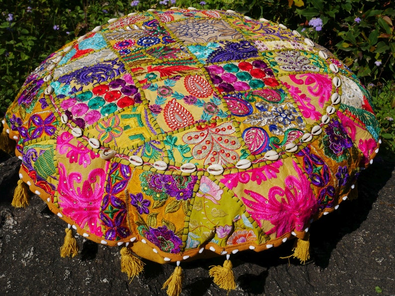 May include: A round, multi-coloured floor cushion with a patchwork design. The cushion is made of fabric with various embroidered floral patterns and is decorated with white seashells around the edge. The cushion has yellow tassels around the bottom.