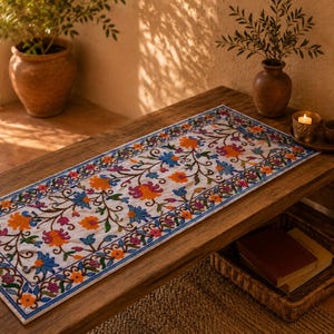 Peut inclure: Un chemin de table rectangulaire blanc orné d'un motif floral brodé complexe dans des tons vifs d'orange, de bleu, de rose et de vert. Le design se compose de vignes, de feuilles et de fleurs entrelacées disposées en une bordure continue.