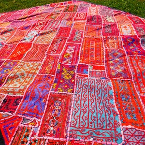 Può includere: Un vivace tessuto patchwork con una varietà di quadrati di tessuto fantasia nei toni del rosso, rosa, arancione e viola. I quadrati sono bordati da cuciture bianche e incorniciati da un bordo nero. Il tessuto è appoggiato sull'erba verde.