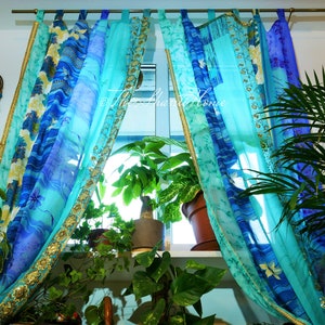 Op de afbeelding: Een set van transparante gordijnen in een blauw en turquoise kleurenschema. De gordijnen hebben een bloemenpatroon en gouden bies. De gordijnen hangen in een raam met planten ervoor.