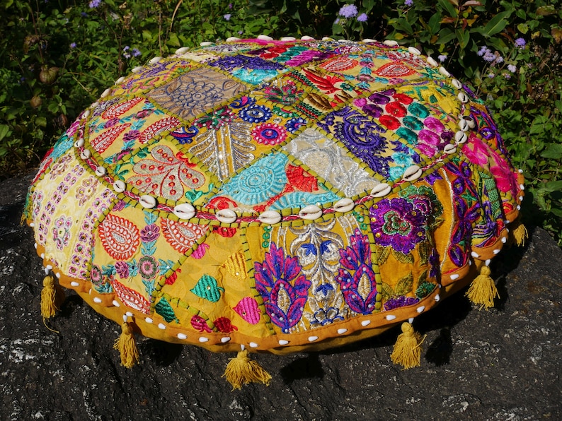 May include: A round, yellow, patchwork floor cushion with colourful embroidery and white shell accents. The cushion has tassels around the edge.