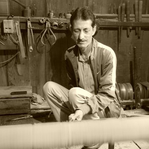 May include: A sepia-toned image of a man working in a workshop. He is seated, focused on a spool of thread or yarn. The workshop has wooden walls and various tools hanging on the wall.